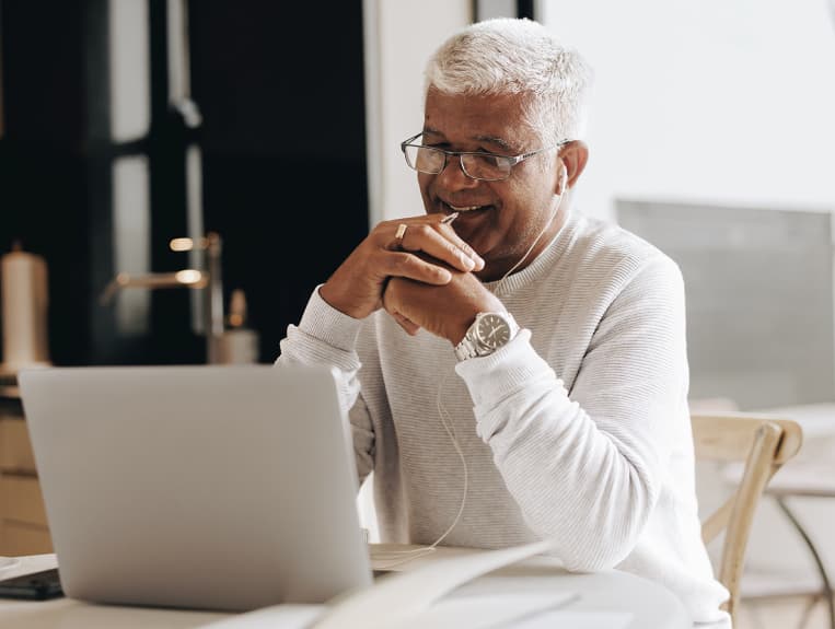 Man smiling looking at a laptop screen and listening to earbuds with hands clasped casually holding a pen.
